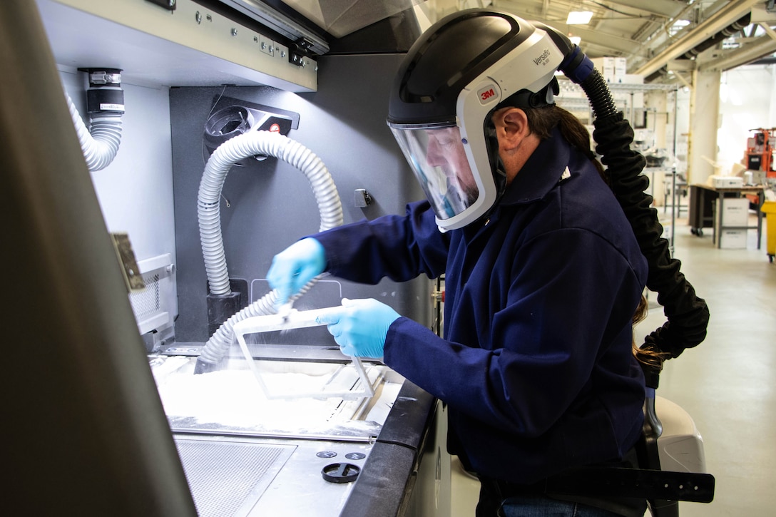 A man wearing protective equipment that includes a helmet with a hose attached to its back works at a manufacturing work station.