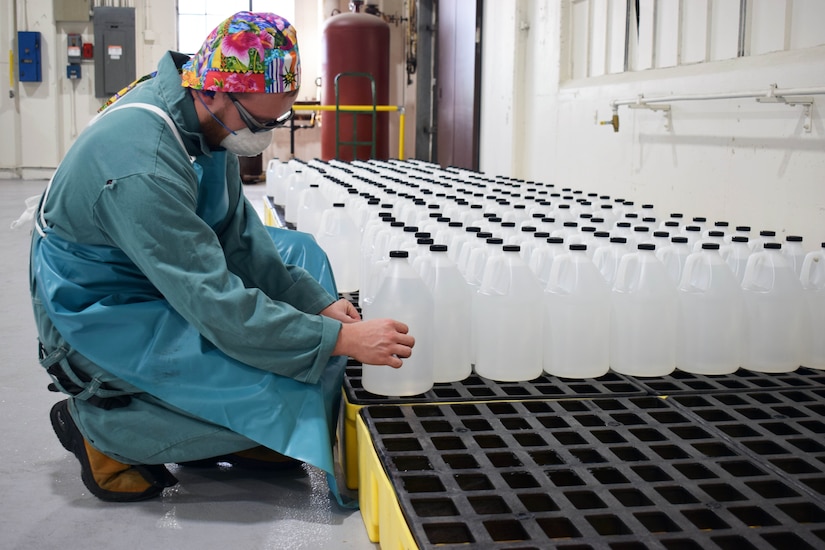 A man wearing protective equipment affixes a label to one of dozens of plastic gallon jugs on pallets.