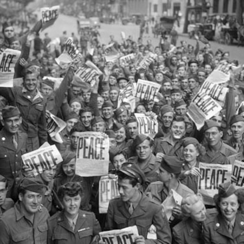 A crowd of people on a city street smile and hold up newspapers with headlines that say "PEACE" in big letters.