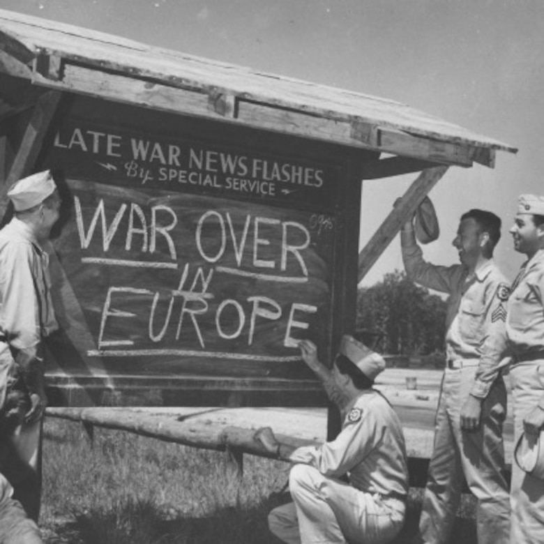 Smiling troops gather round a chalkboard posted outside with "WAR OVER IN EUROPE" written on it.