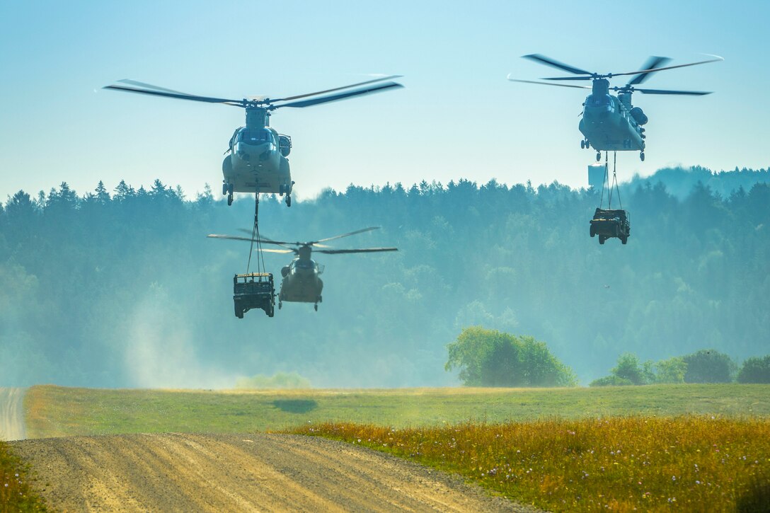 Three Army helicopters fly over rolling terrain, with vehicles suspended by sling from two of them.