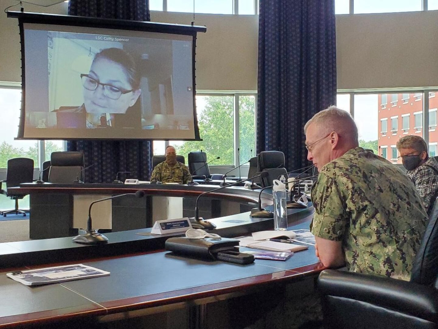 Rear Adm. Grafton Chase is seated at a conference table addressing a virtual participant projected onto a screen