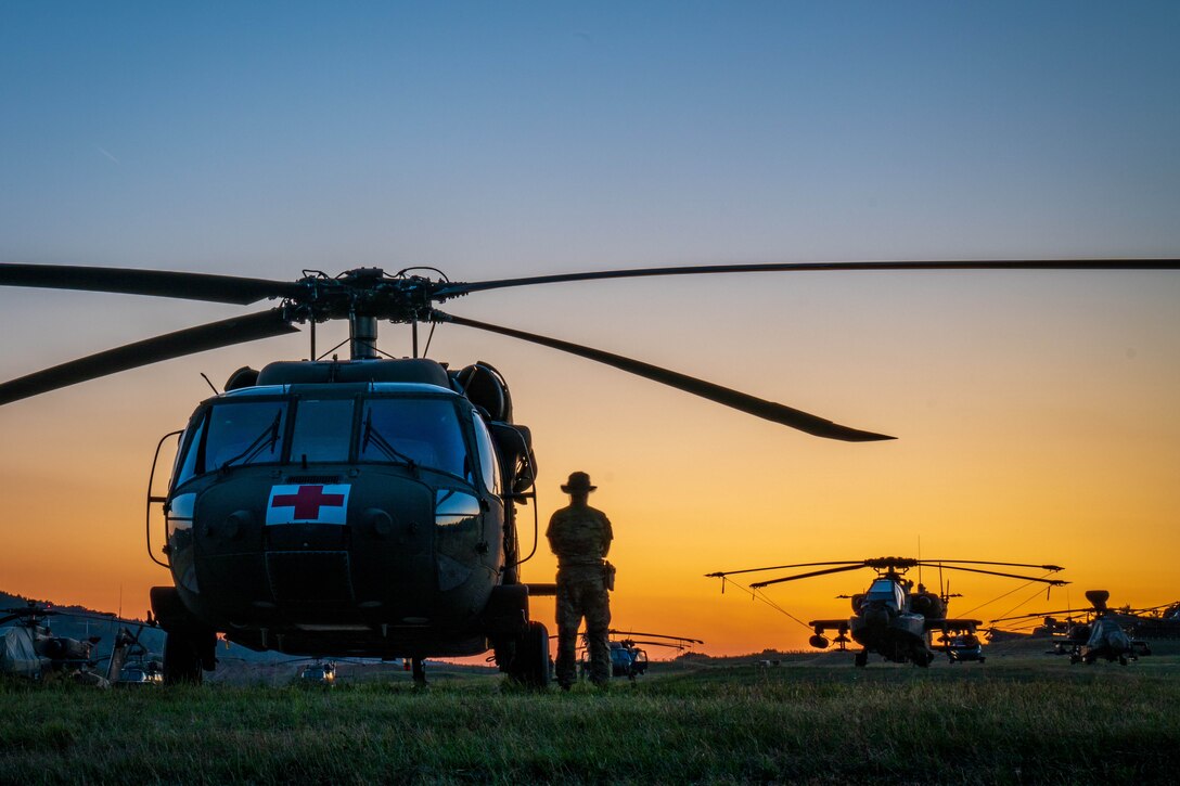 A soldier stands next to a helicopter with others in the background at sunset.