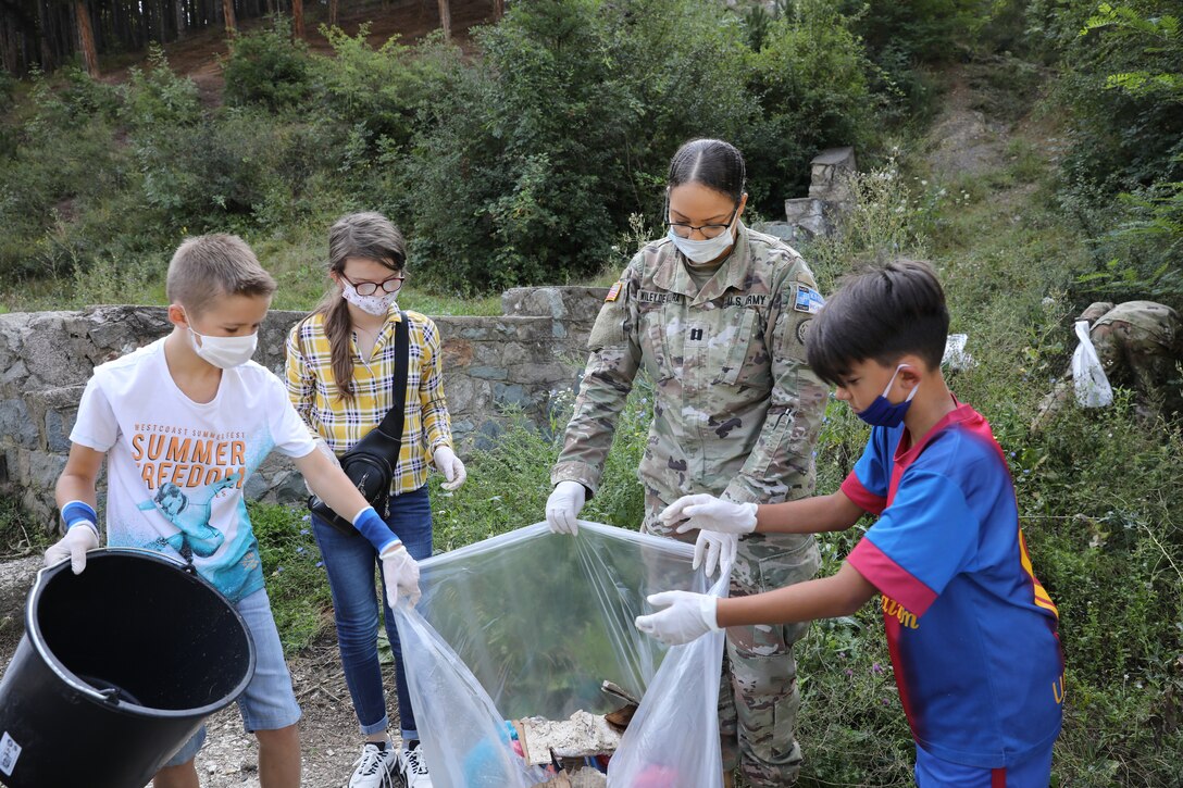 A soldier helps to hold a large trash bag as kids put trash into it.