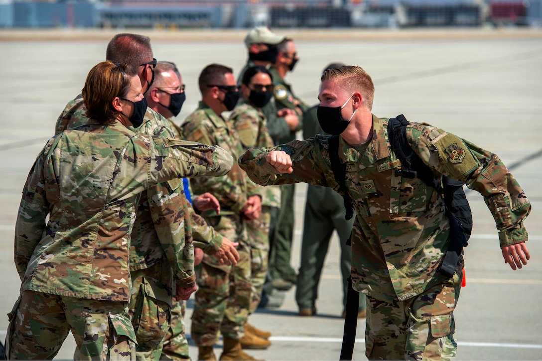 Air National Guard airmen wearing face masks greet each other by bumping elbows while several other airmen wearing face masks watch.