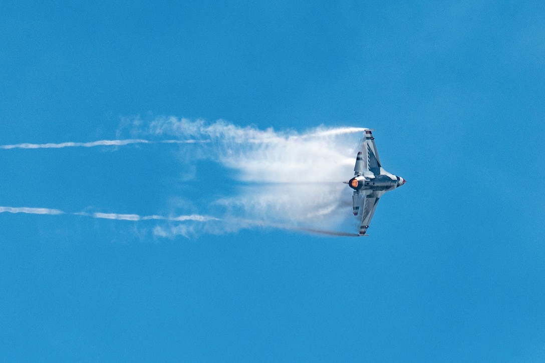 An aircraft flies sideways through a blue sky leaving a white trails in its wake.