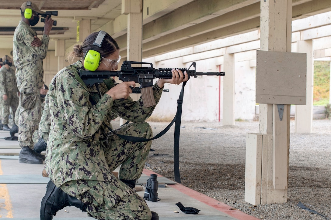 A sailor kneels and fires a weapon while wearing protective ear coverings at a range.