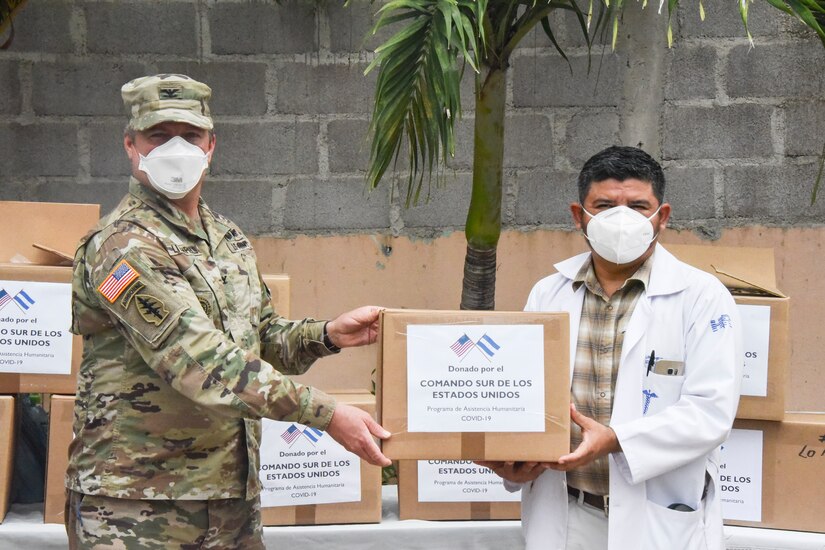 A soldier and a civilian, both wearing face masks, pose for a photo as they hold a box.