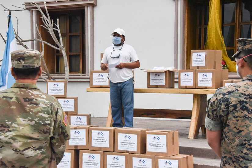 A man wearing a face mask, a white baseball cap, a T-shirt and jeans stands amid boxes of humanitarian aid supplies and speaks to the soldiers who delivered the items.