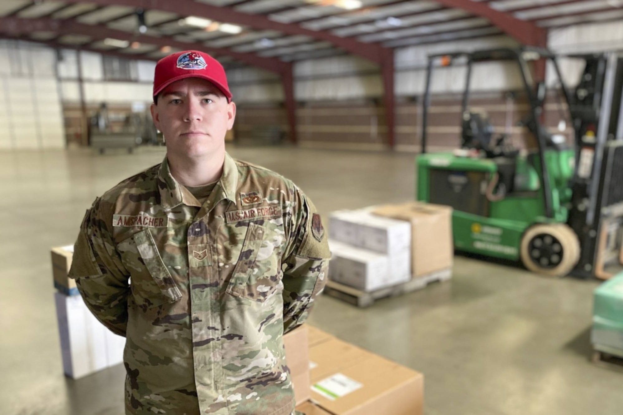 An Airmen wearing a red hat stands at parade rest in a warehouse