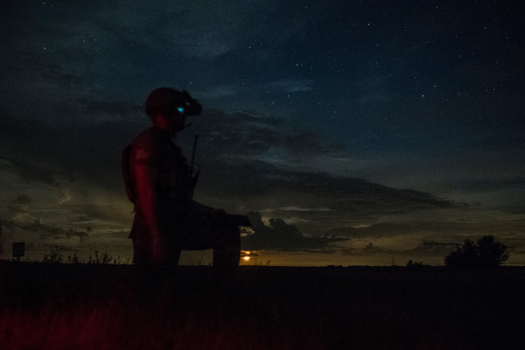 A single special operations troop kneels on one knee in front of a rapidly fading sunset. The troop is using night vision equipment as the light fades.