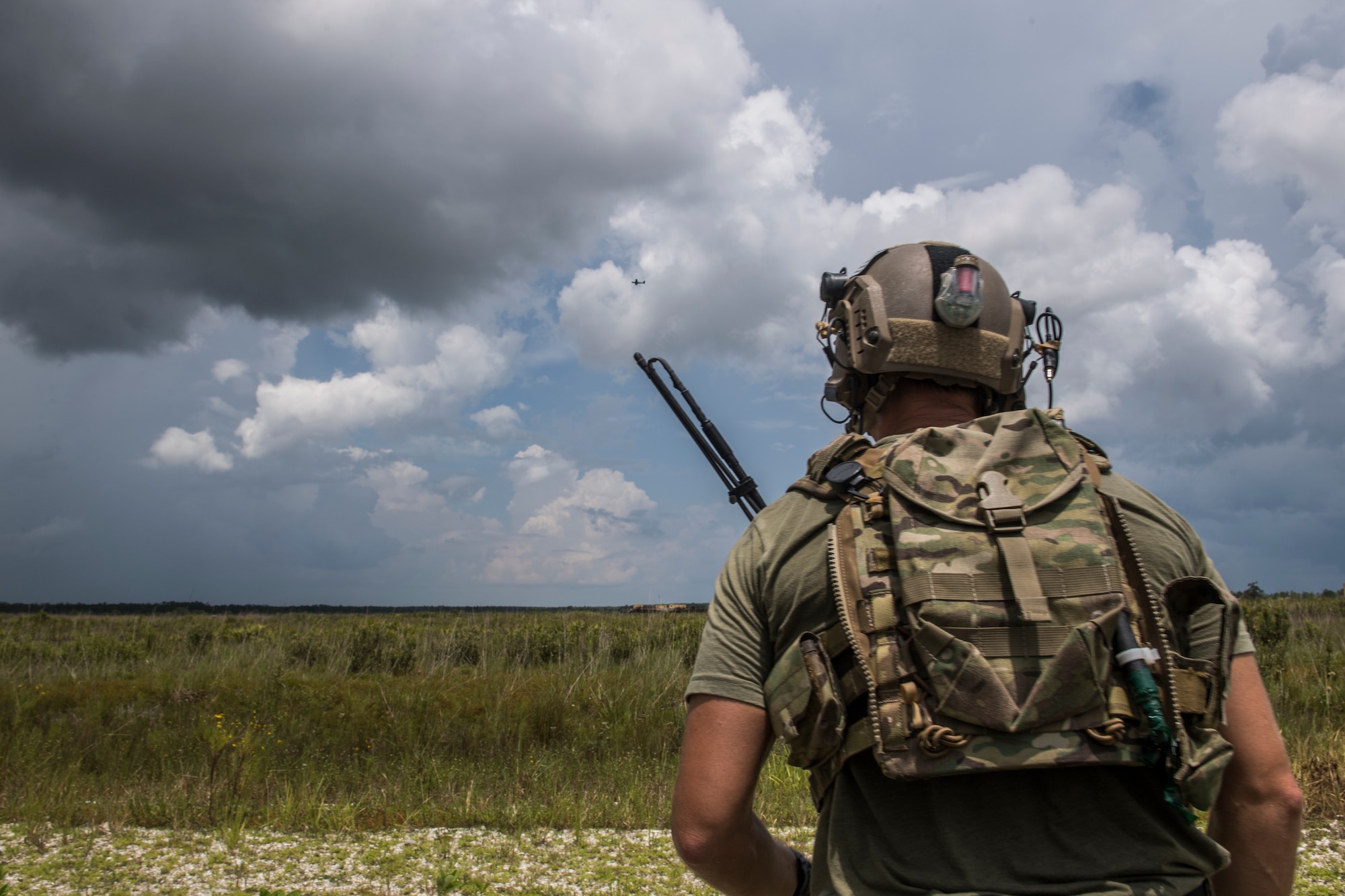 A special operations troop stands with his backpack to the camera looking over a landing area of sand and tall grass with mixed clouds in a blue sky in the background.