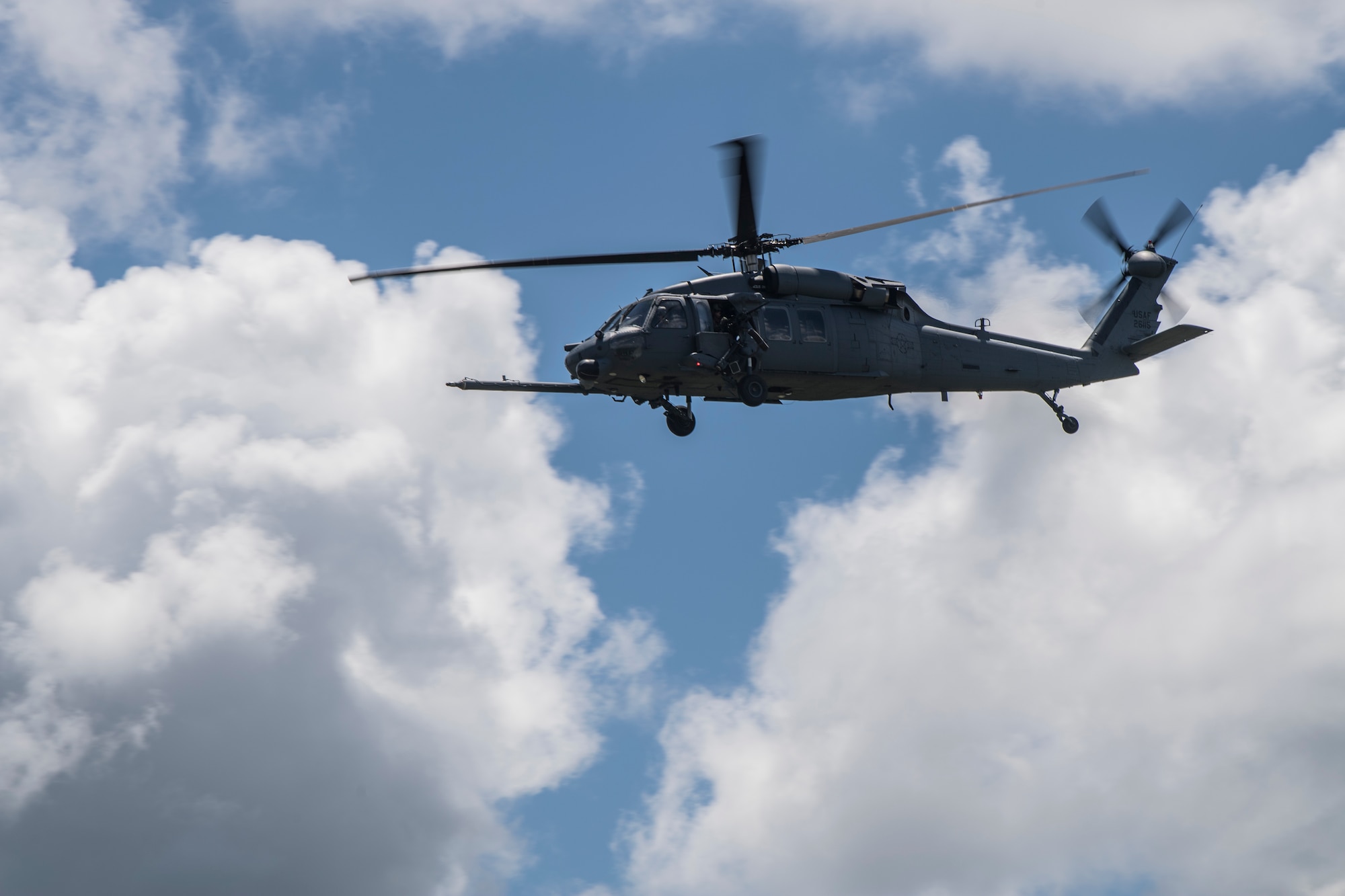 An HH-60 Pave Hawk helicopter hovers in side profile against a backdrop of white clouds in a blue sky.