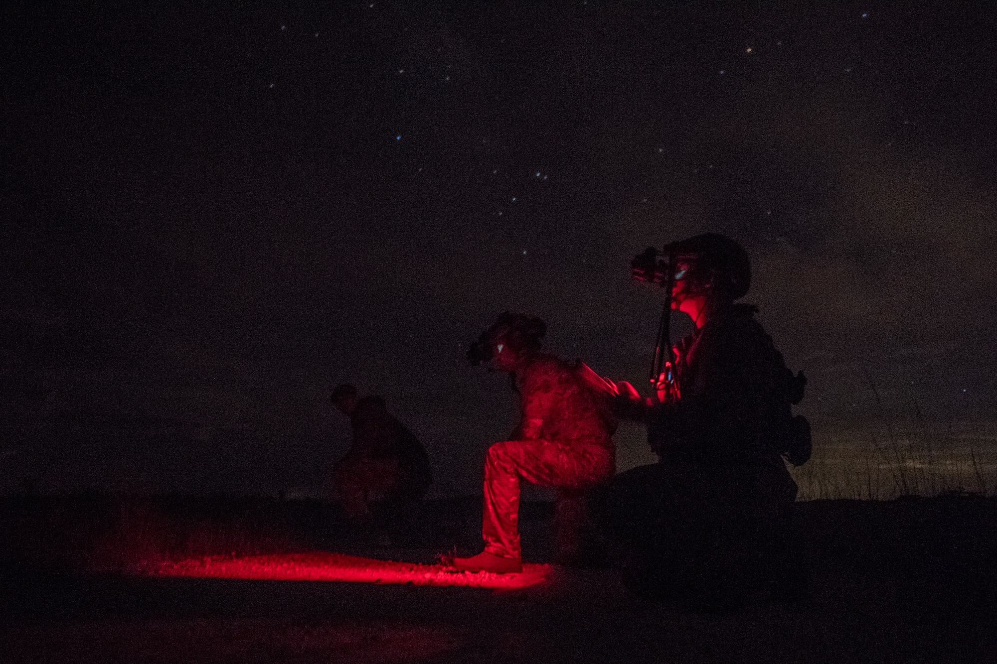 A pair of special operations troops kneel on one knee at night, lit only from low intensity red lighting. Special operations troops from the Army, Air Force, and Marine Corps conduct close air support during the Special Operations Terminal Attack Controller Course at Avon Park Florida, Aug. 6, 2020.