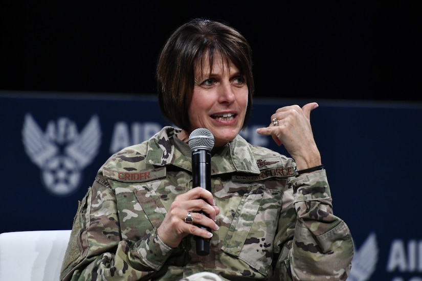 Air Force general gestures with her left hand while speaking to a group and holding a microphone in her right hand.