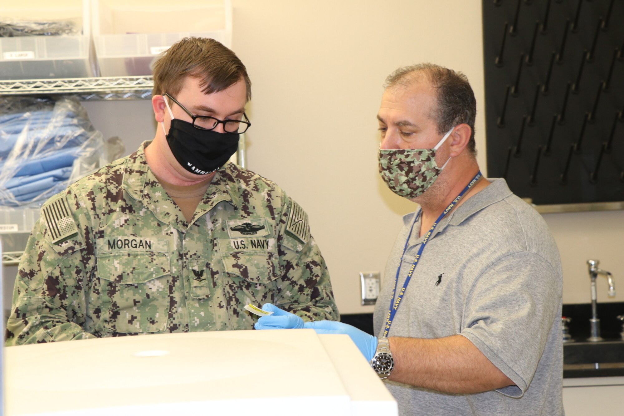 A Navy Corpsman in uniform listens to a civilian employee speak in a hospital lab.