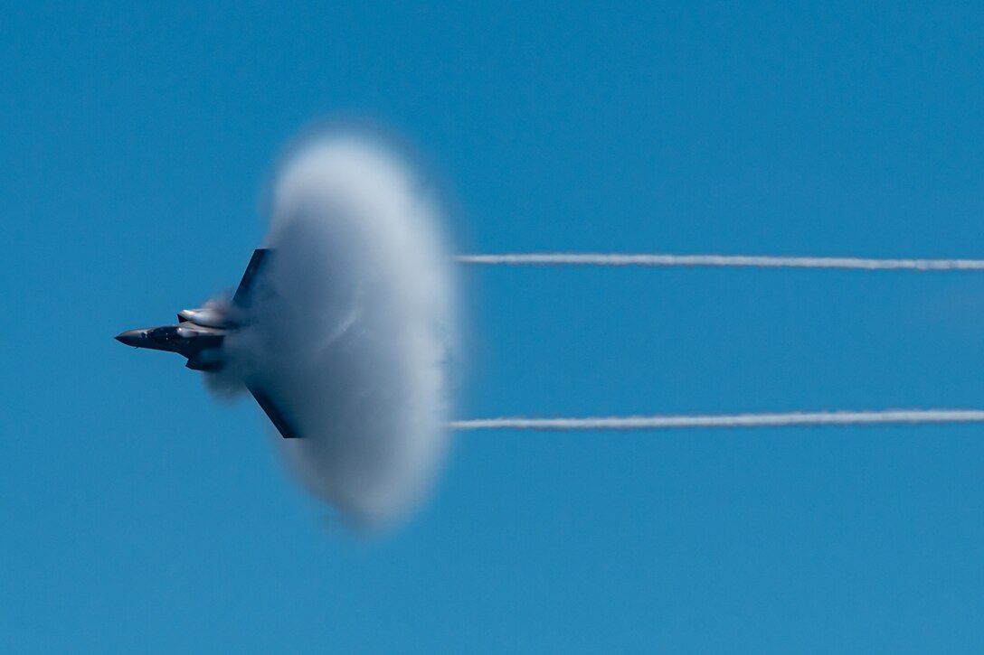 A military jet emits a large puff of smoke as it flies against a blue sky,