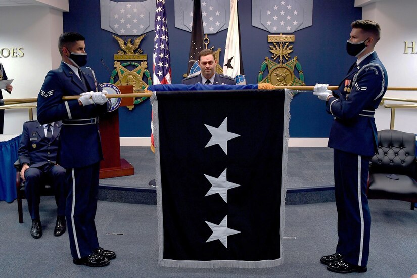 Two service members hold a flag between them as another man stands behind it.