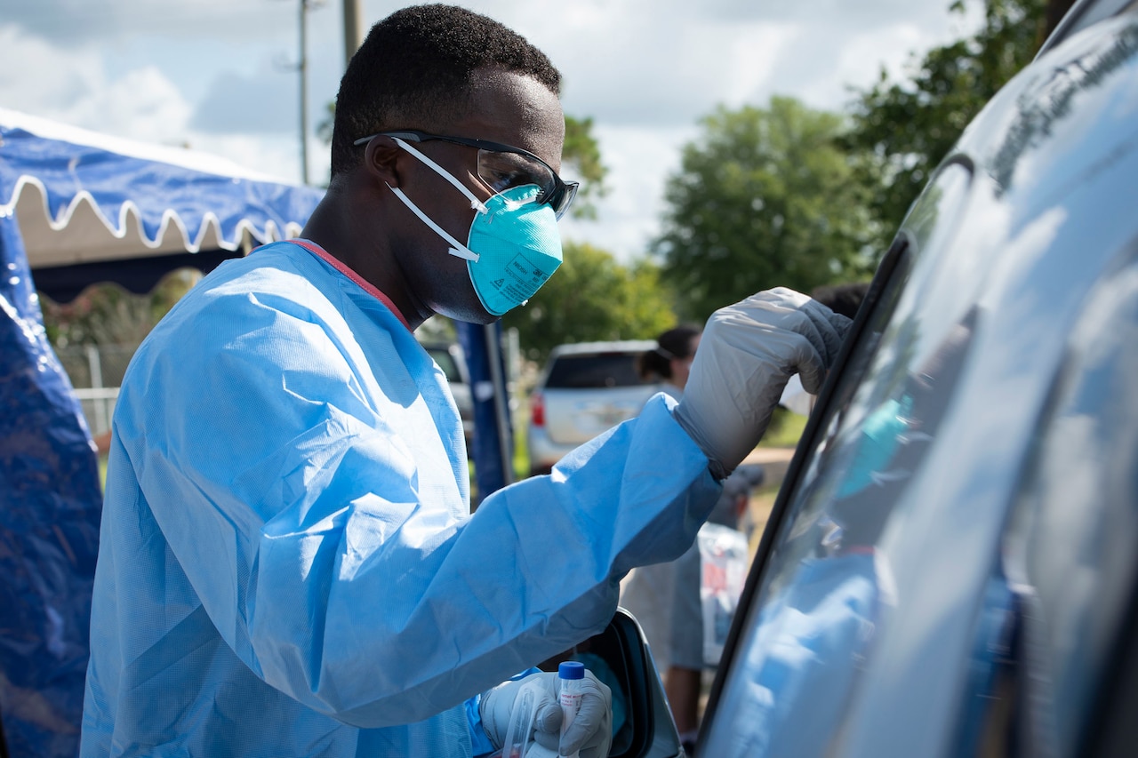 An airman wearing personal protective equipment administers a COVID-19 test to a person in their car.