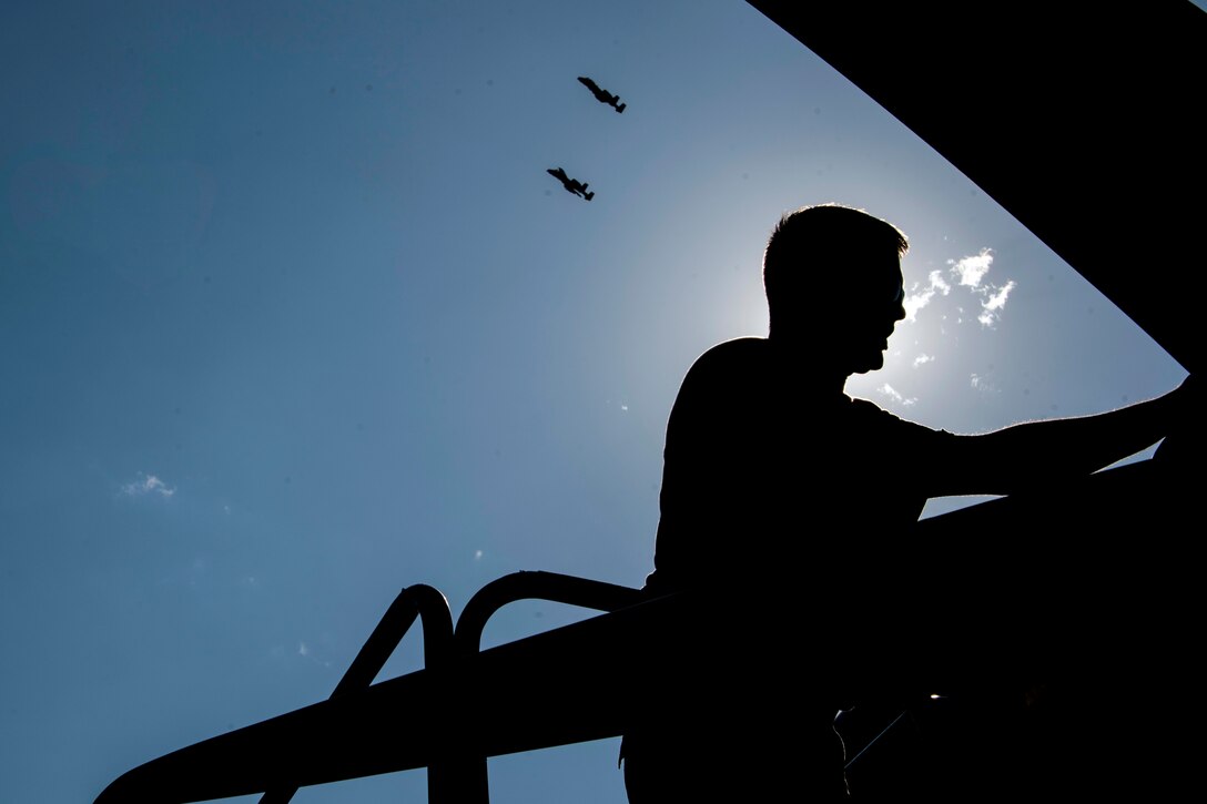 An airman, shown in silhouette, works on an aircraft as two others fly in the background.