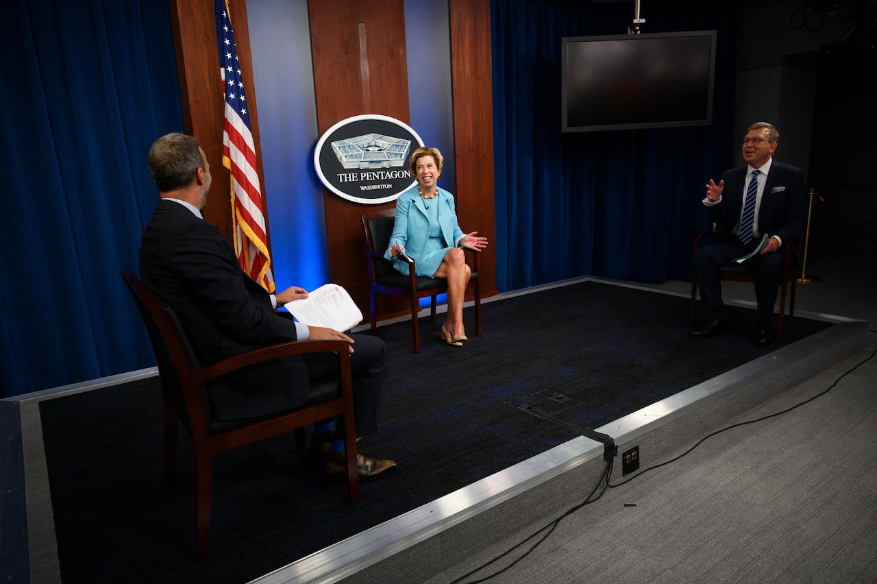 Three individuals sit in chairs arranged on a platform. A sign on the wall reads “The Pentagon -- Washington.”