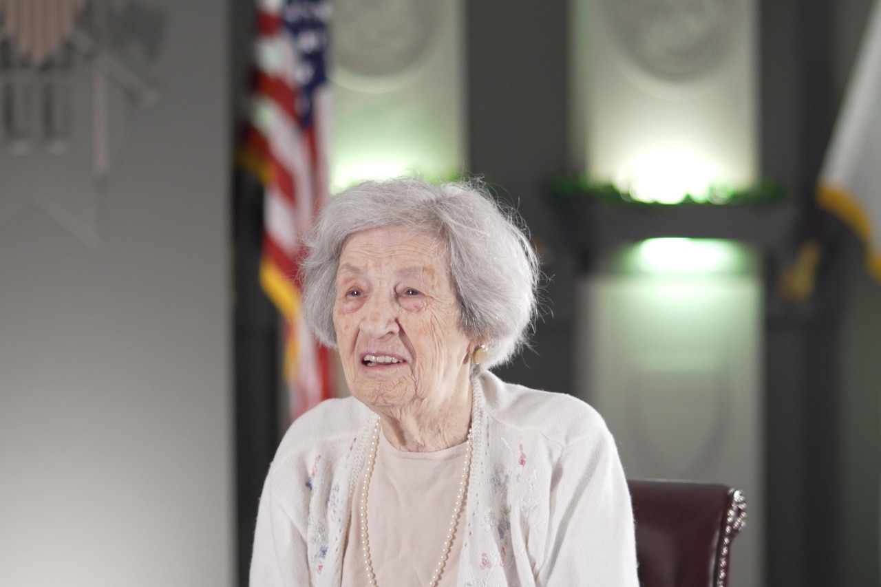 A woman sits in a chair flanked by the U.S. and U.S. Army flags.