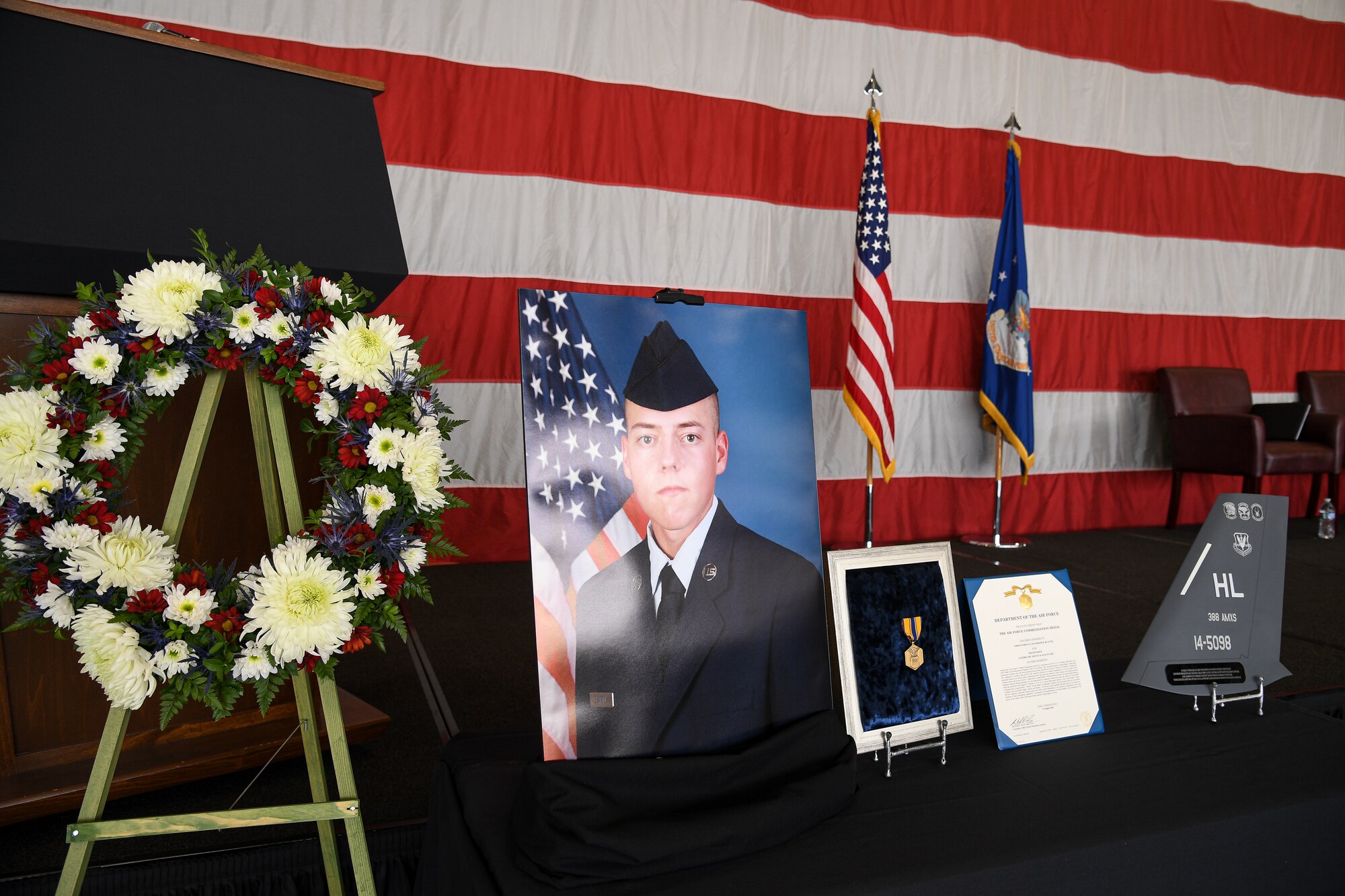 A table sits in front of an American flag with photos, a medal, and a wreath for a fallen Airman.
