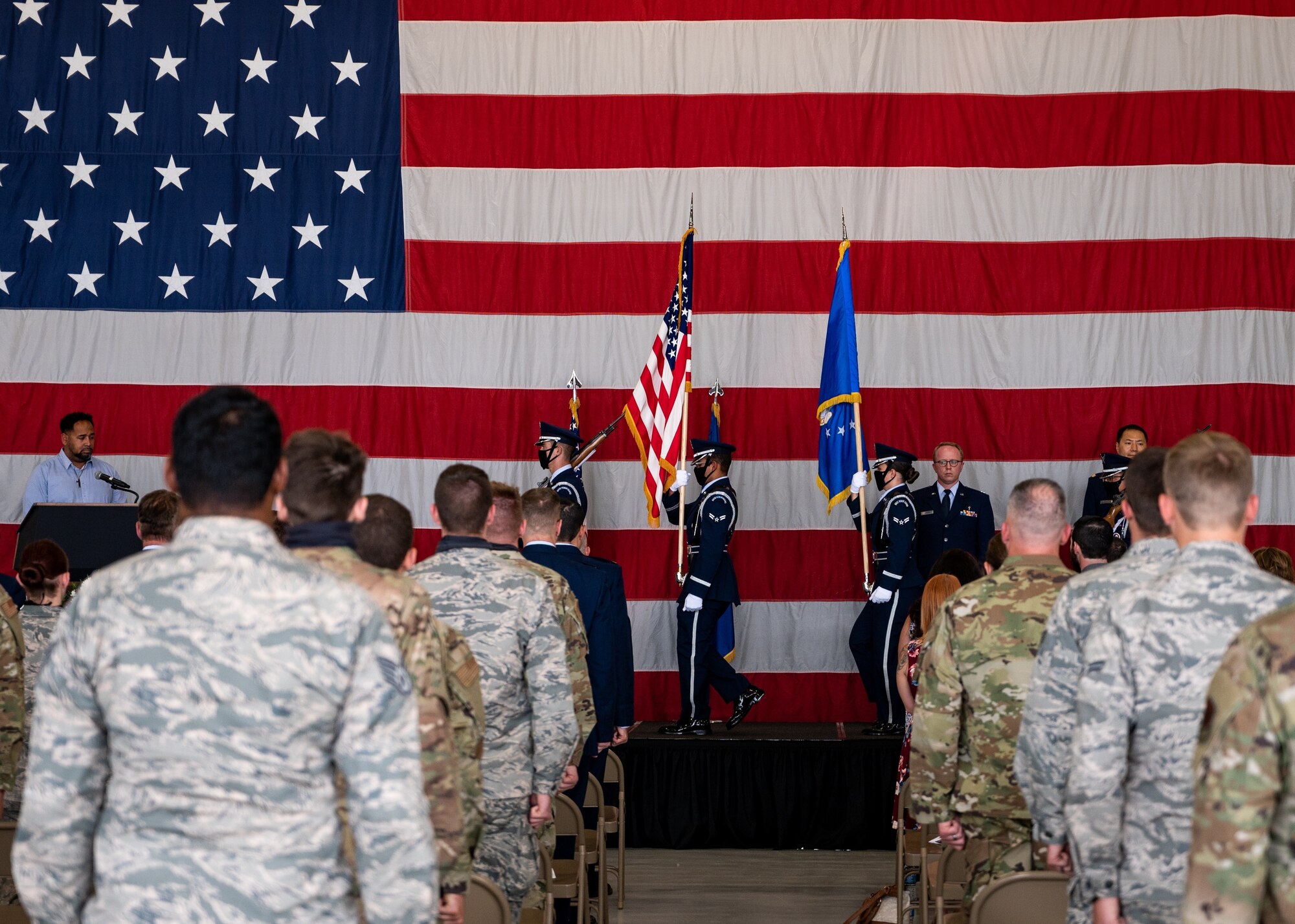 Uniformed personnel stand in from an American flag while a U.S. Air Force color guard details marches on a stage.