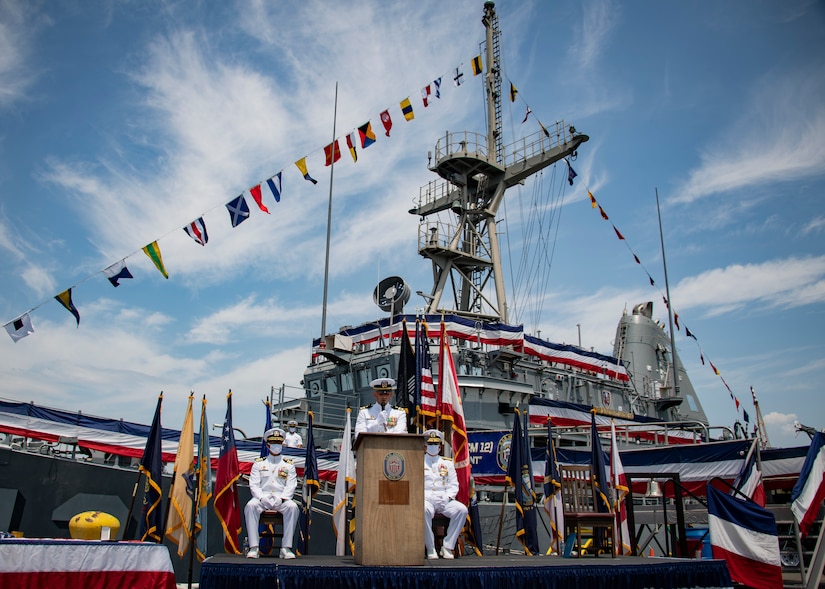 Sailors in uniform sit in front of a ship during a decomissioning ceremony.
