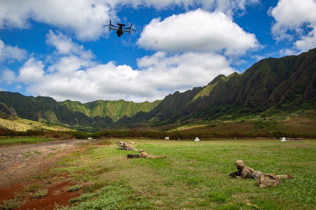 Marines lay on the ground near mountains as an aircraft flies over.