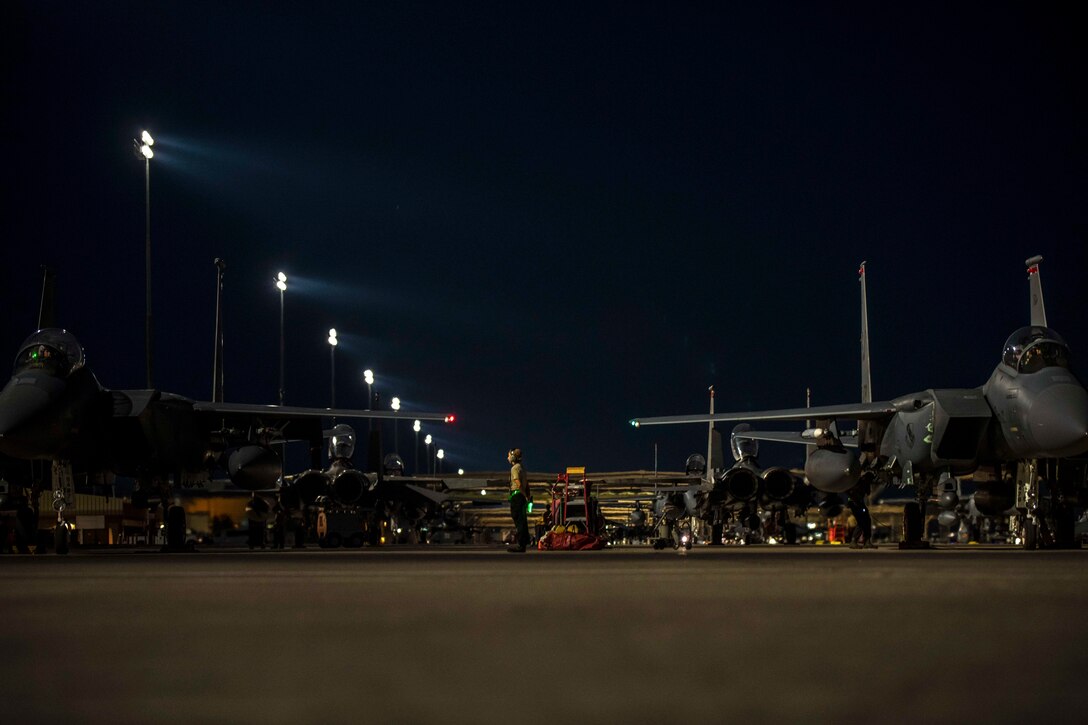 An airman stands between several aircraft in the dark.