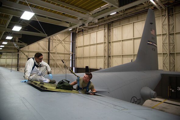 Two Airman sit on top of a C-130 wing inside of a hangar and connect electrical components inside of a panel.