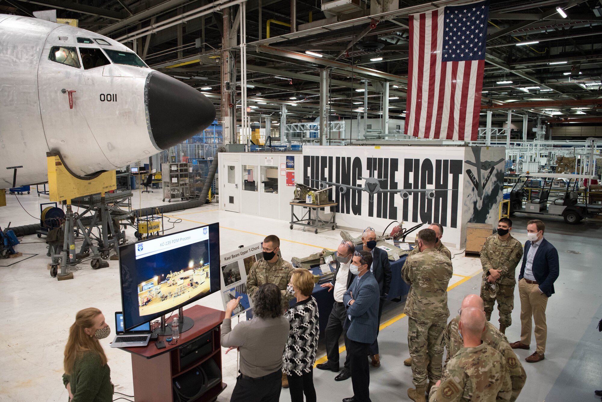 Group of Airmen and Civilians looking at TV with aircraft undergoing maintenance in background.