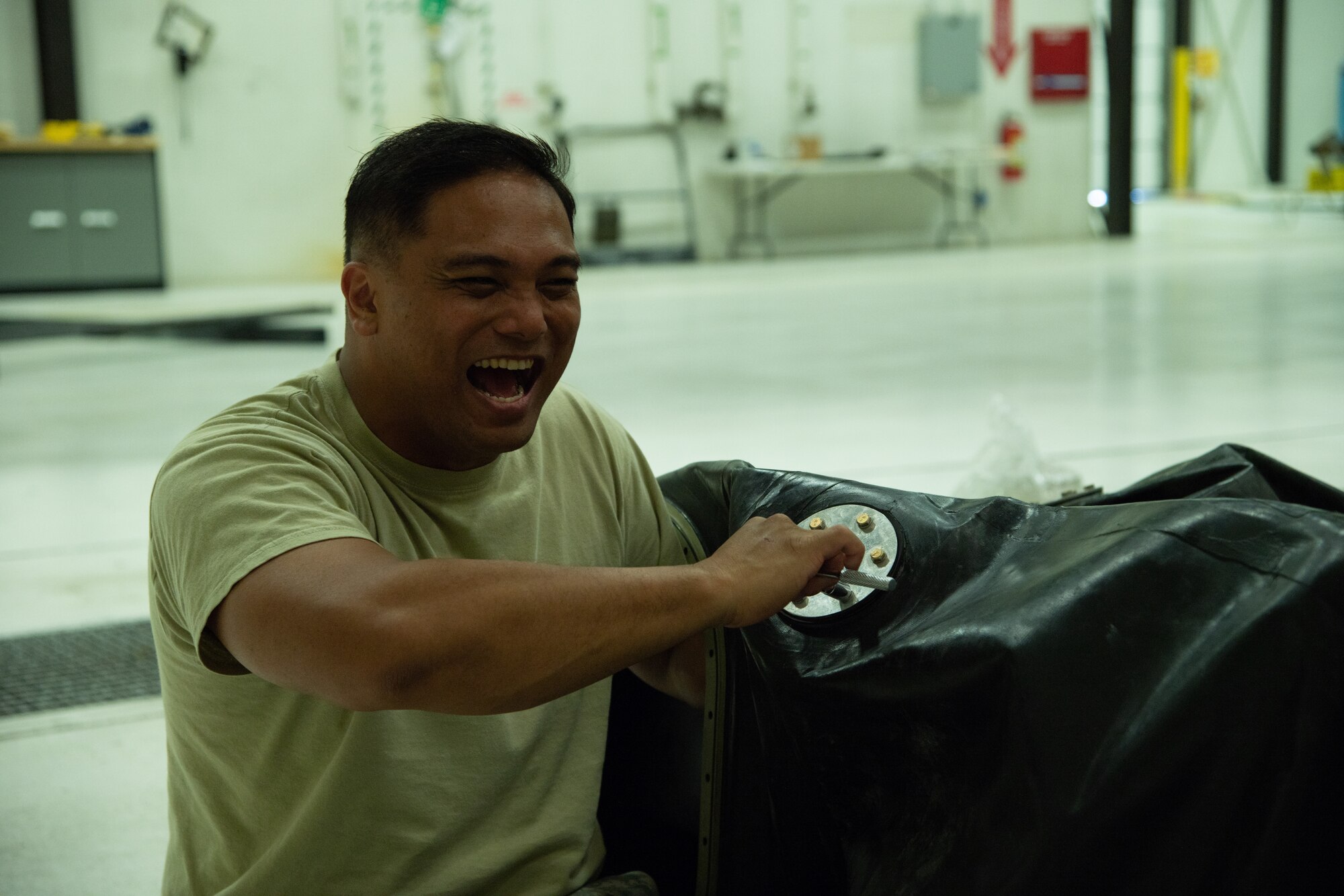 Fuel system technician uses wrench to screw cover plate onto large black rubber fuel cell while joking with his team.