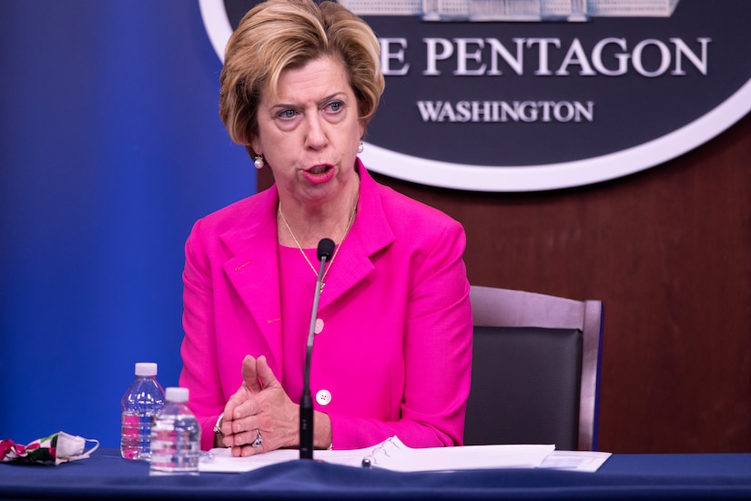 A woman sits at a table. Behind her is a sign that reads “The Pentagon - Washington.”