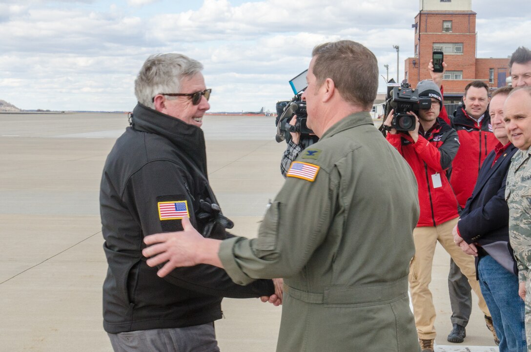 Two men shake hands on an airfield as cameras in the background record the interaction.