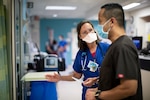 Woman in blue scrubs and white face mask talks with a  man in black scrubs and blue face mask in a room with medical equipment.