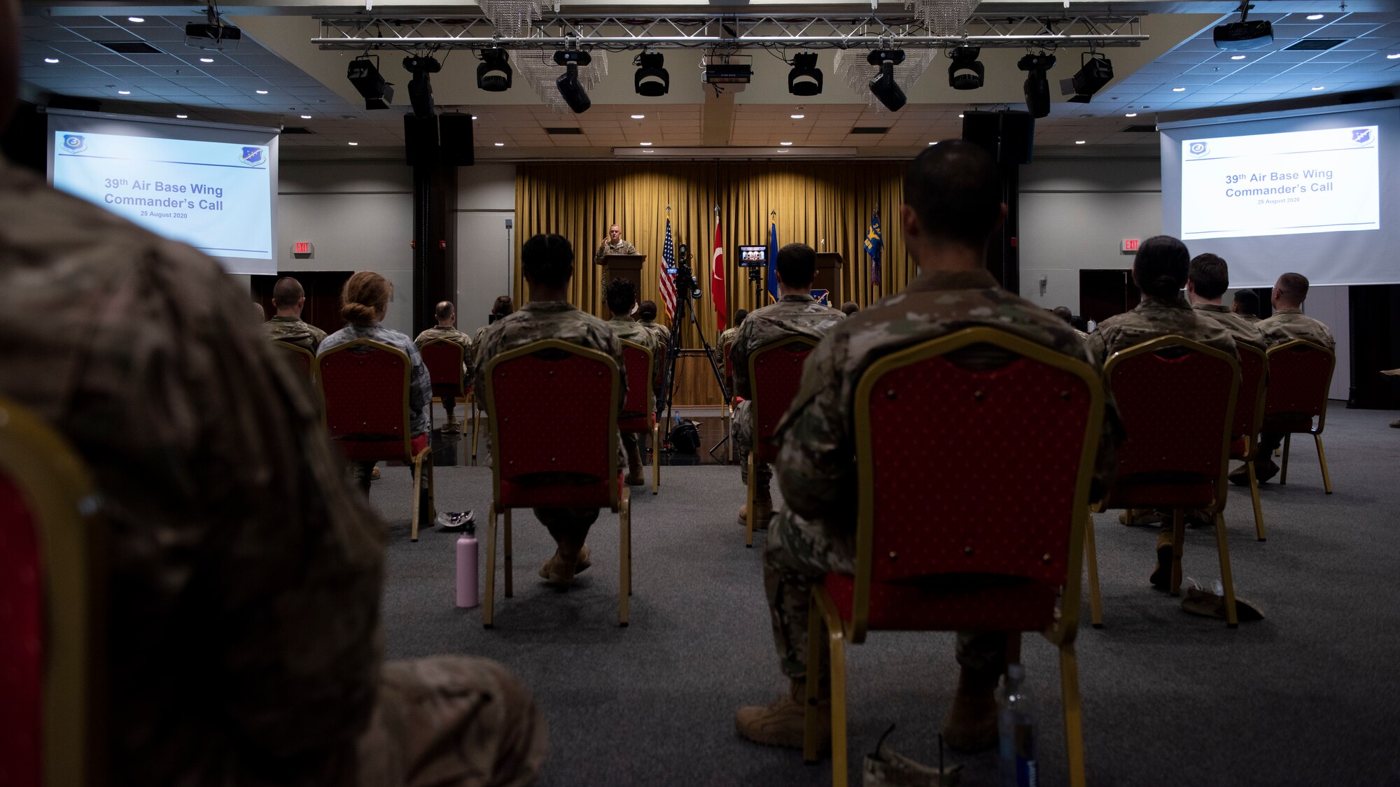 Airmen attending a commander's call meeting sit spaced out six feet each while the base commander speaks from stage in the back of the room, August 25, 2020.