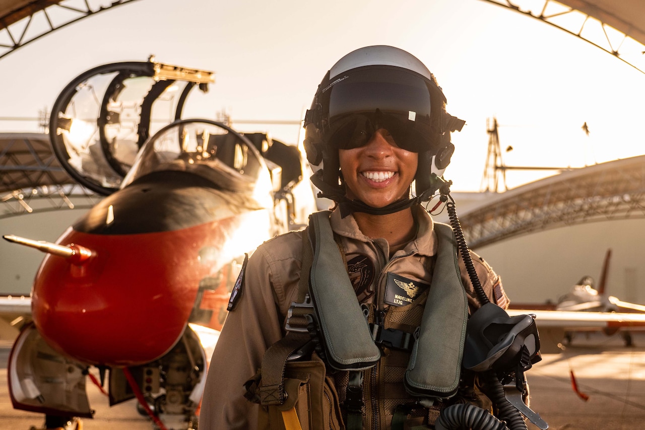 A pilot with a helmet on stands in front of a jet near a hangar.