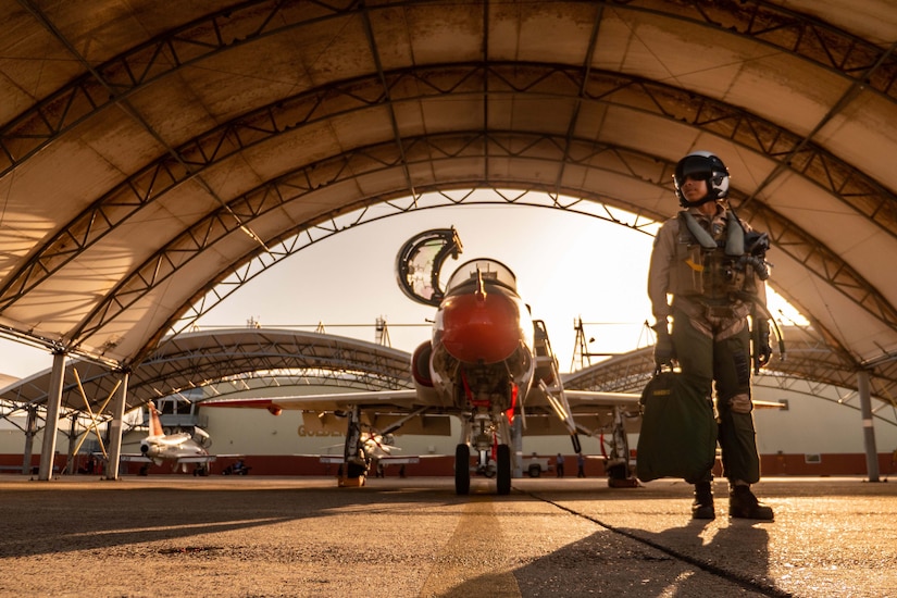 A pilot in combat gear stands beside a jet in a hangar.