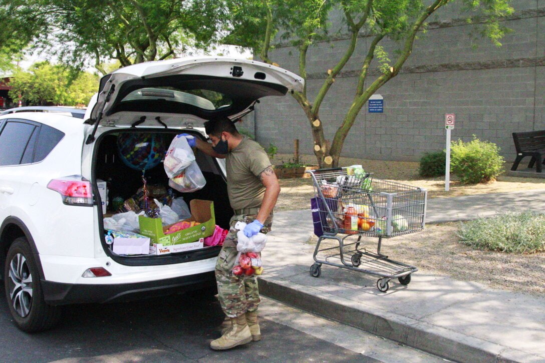 A male service member wearing a face mask puts groceries into the trunk of a car.