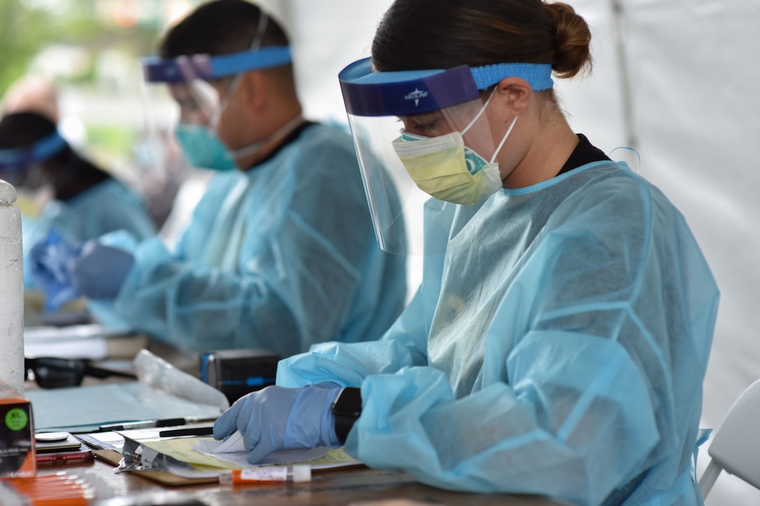 An Army soldier wearing personal protective equipment works at a testing site along with several other soldiers wearing personal protective equipment.