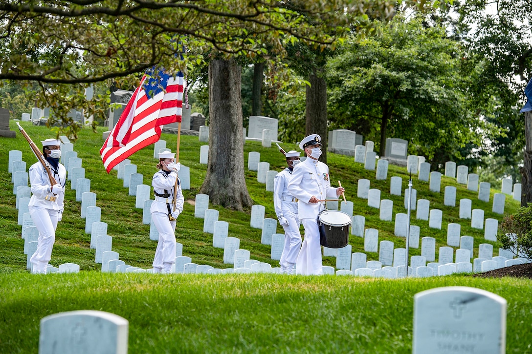 Sailors, including one holding an American flag, and one playing a drum, walk in a cemetery.