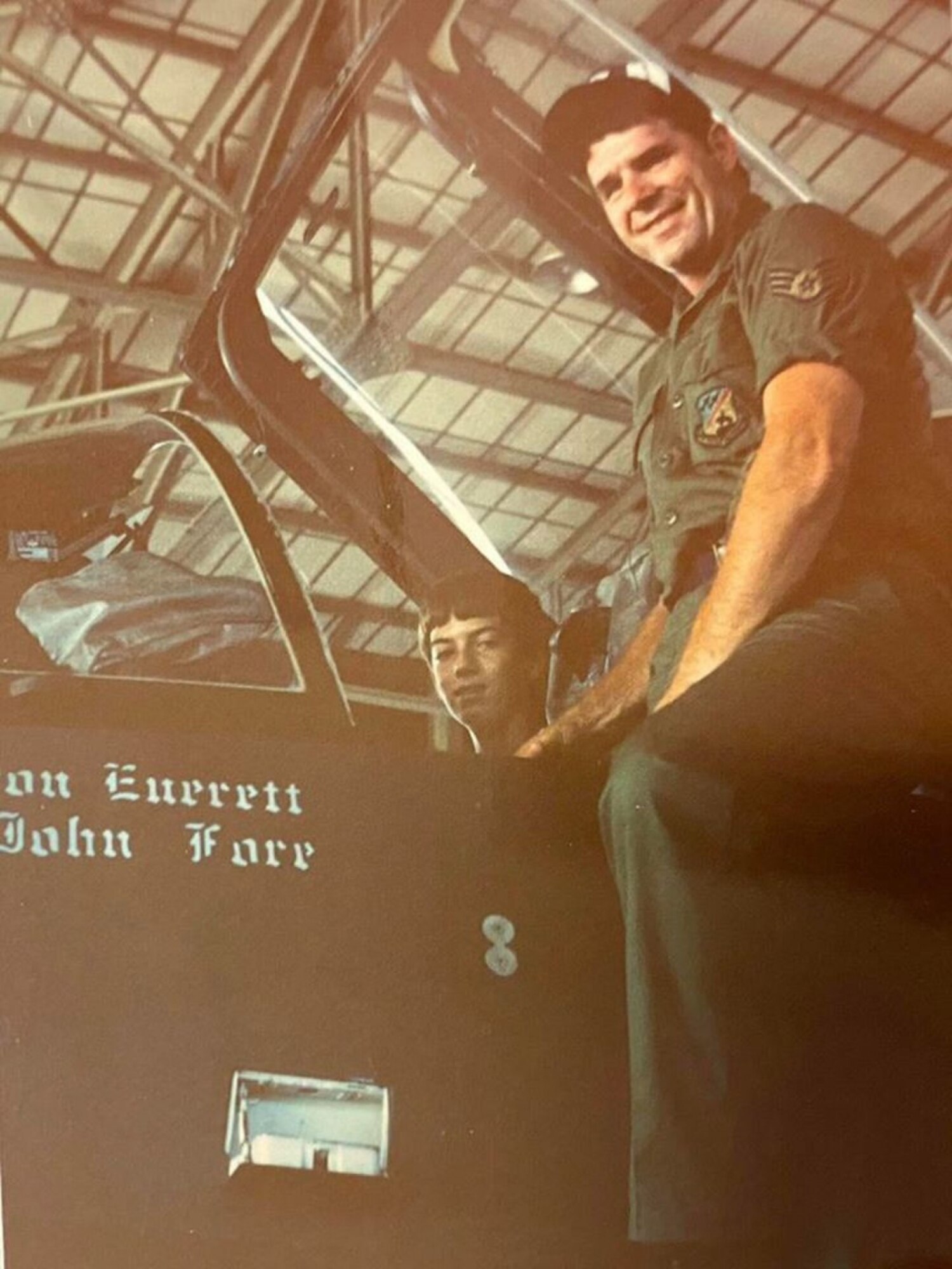 An Airman stands next to the cockpit of a jet while a child sits inside