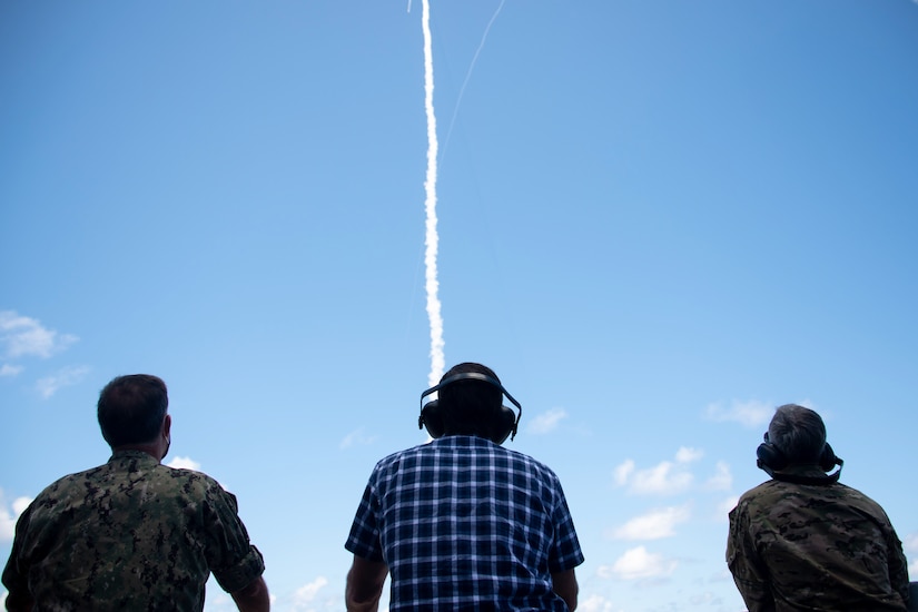 Three people look up at a trail of white smoke in the sky.