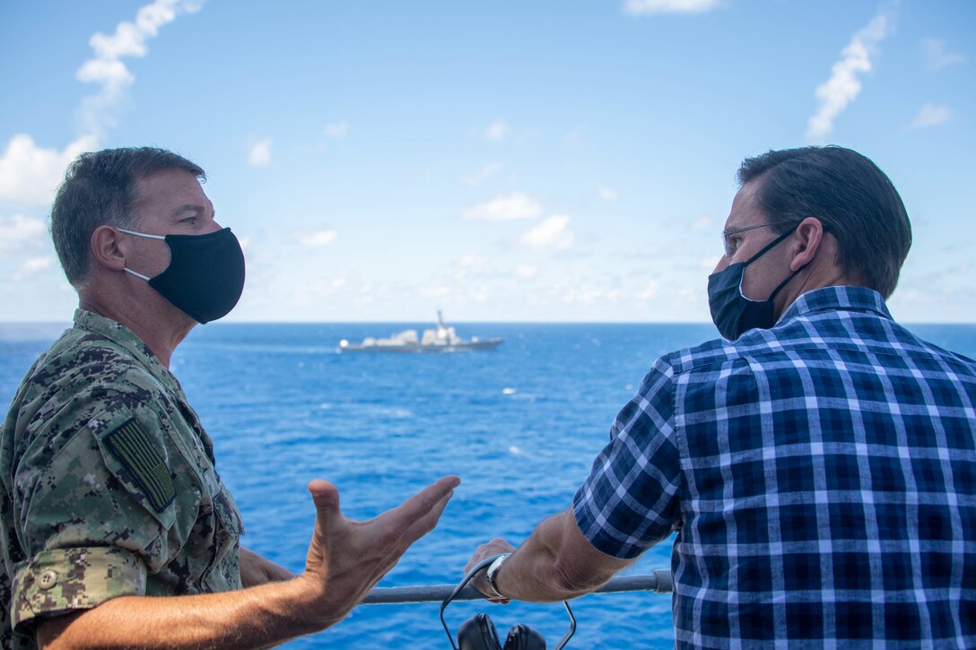 Two men wearing masks speak on a ship deck with the ocean behind them.