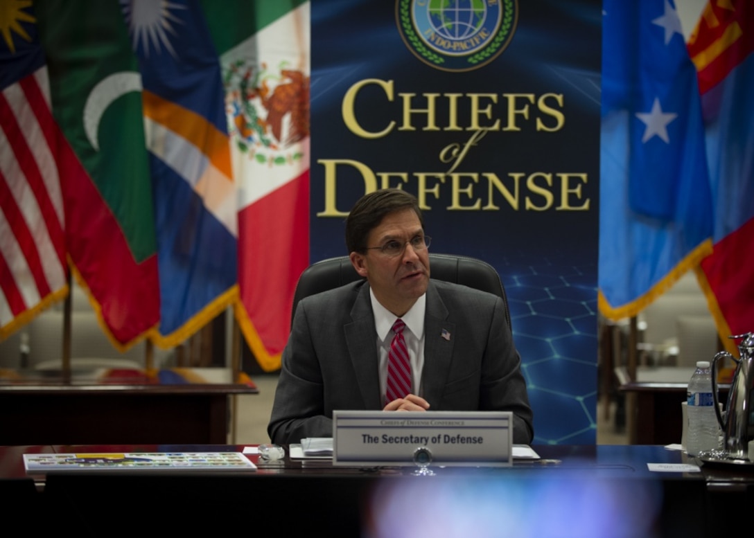 Defense Secretary Dr. Mark T. Esper sits at a table in front a of a Chiefs of Defense sign flanked by series of flags.