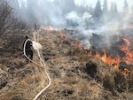 Two people lay hoses across dry landscape with fires and smoke in the distance.