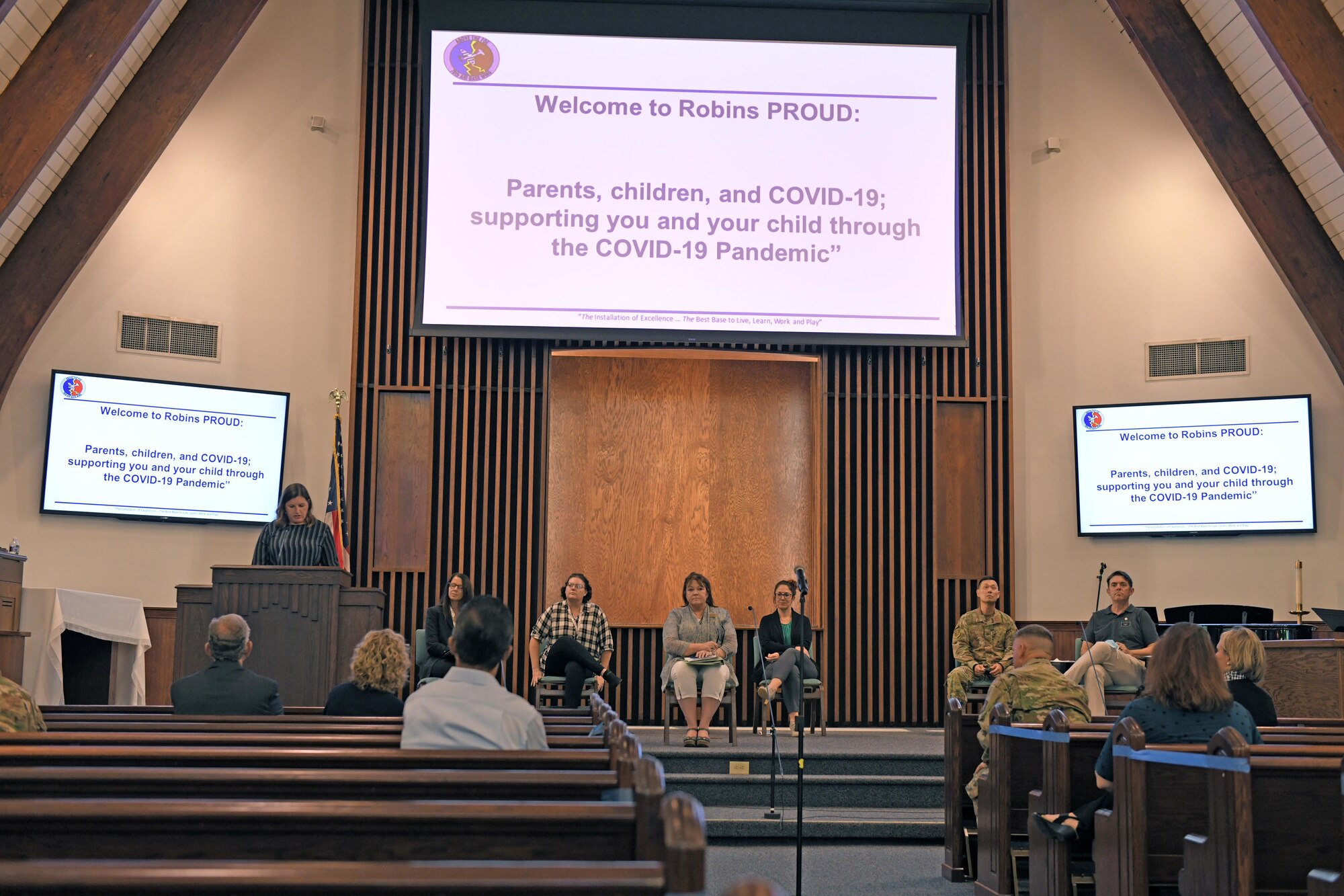 Photo shows panelists sitting on stage with large information screens above them.