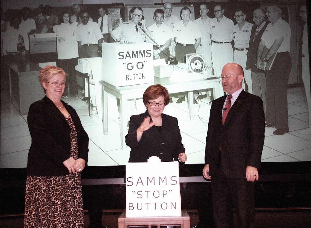 Two women and a man stand in front of a sign that says "SAMMS STOP BUTTON" in front of a historical photo of a group of employees about to hit the "SAMMS GO BUTTON."